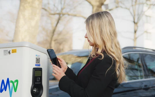 Blonde Frau bedient ein Smartphone an einer Elektroladesäule